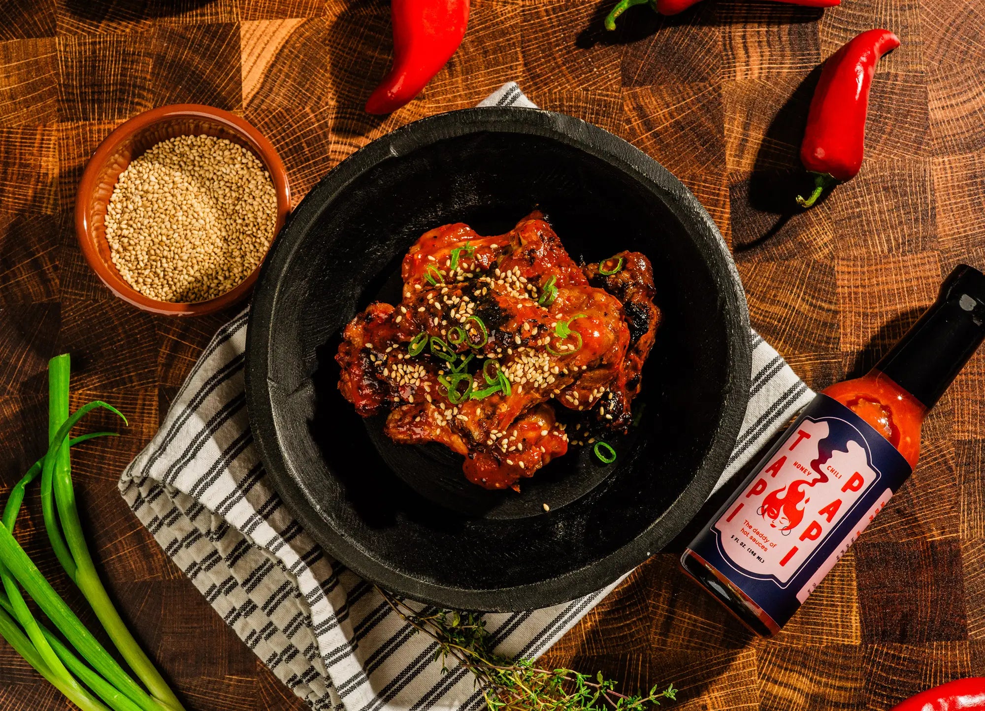 Bowl of chicken wings on a wooden table with spices and a bottle of Tapi Papi hot sauce.
