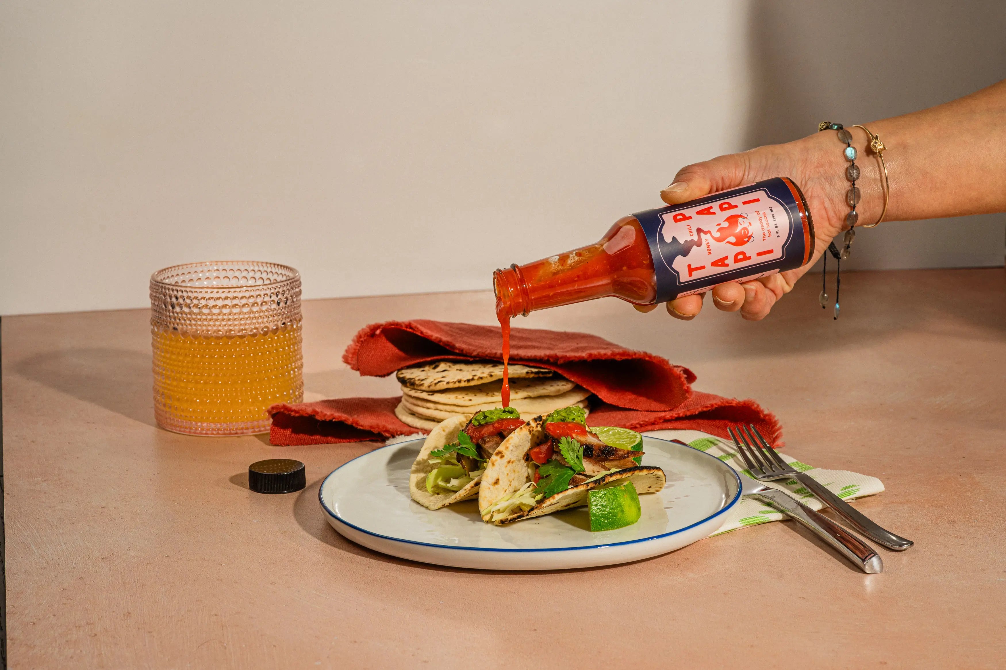 Person pouring Tapi Papi hot sauce over chicken tacos on a table with a glass of beer.