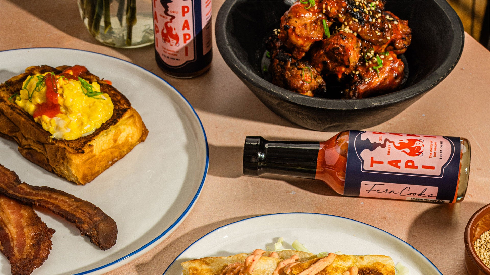 Table with food items including a plate of scrambled eggs on toast, a bowl of chicken wings, and a bottle of Tapi Papi hot sauce.