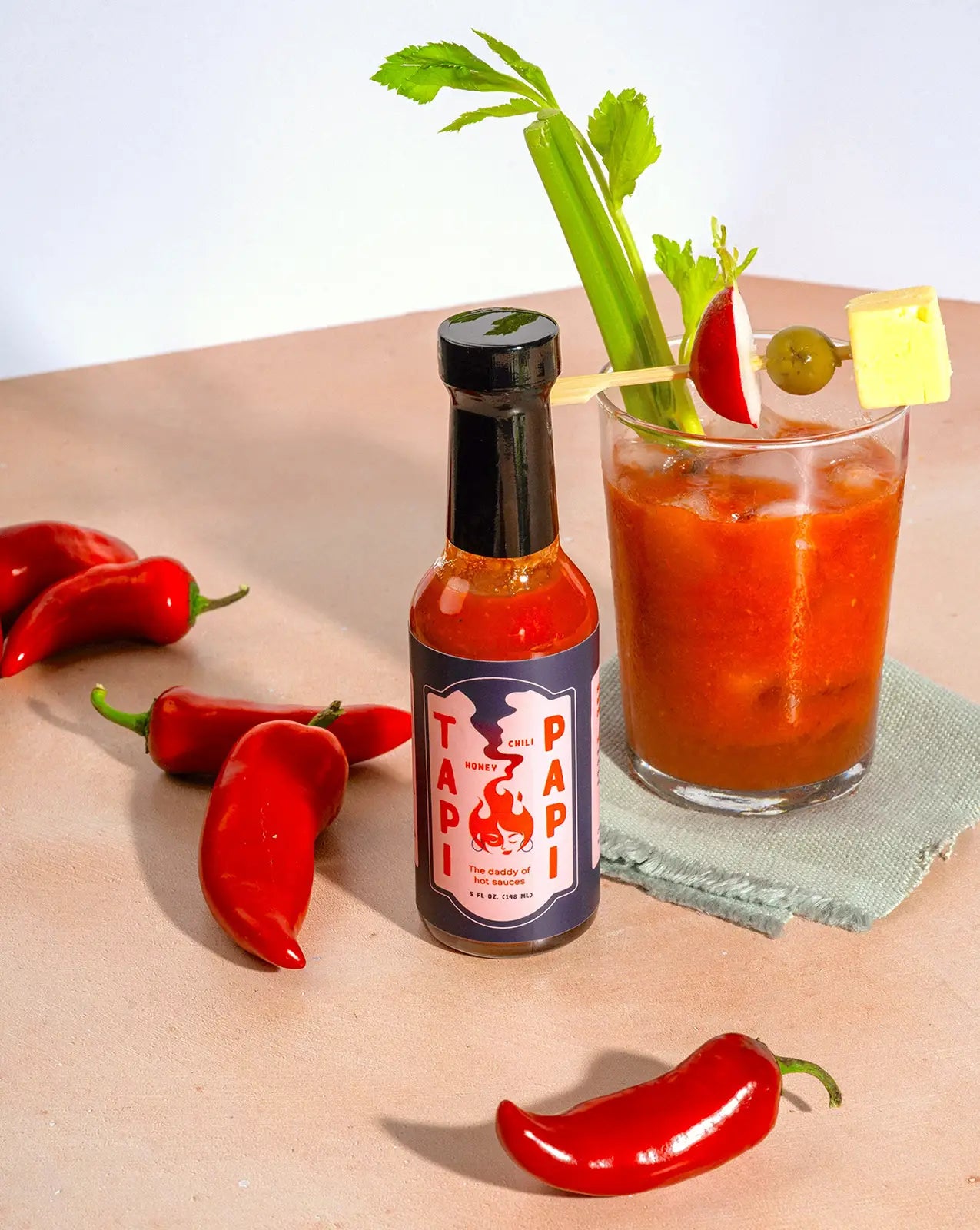 A bottle of Tapi Papi hot sauce with a Bloody Mary and chili peppers on a table.