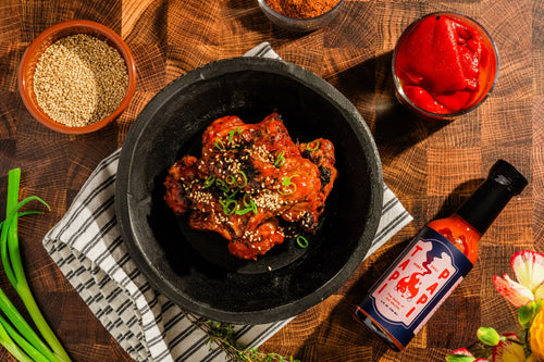Bowl of chicken wings on a wooden table with spices and a bottle of Tapi Papi hot sauce.